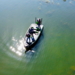 View of fishing boat with three individuals, as seen from above, surrounded by blue-green water with some sunlight glittering near the bow